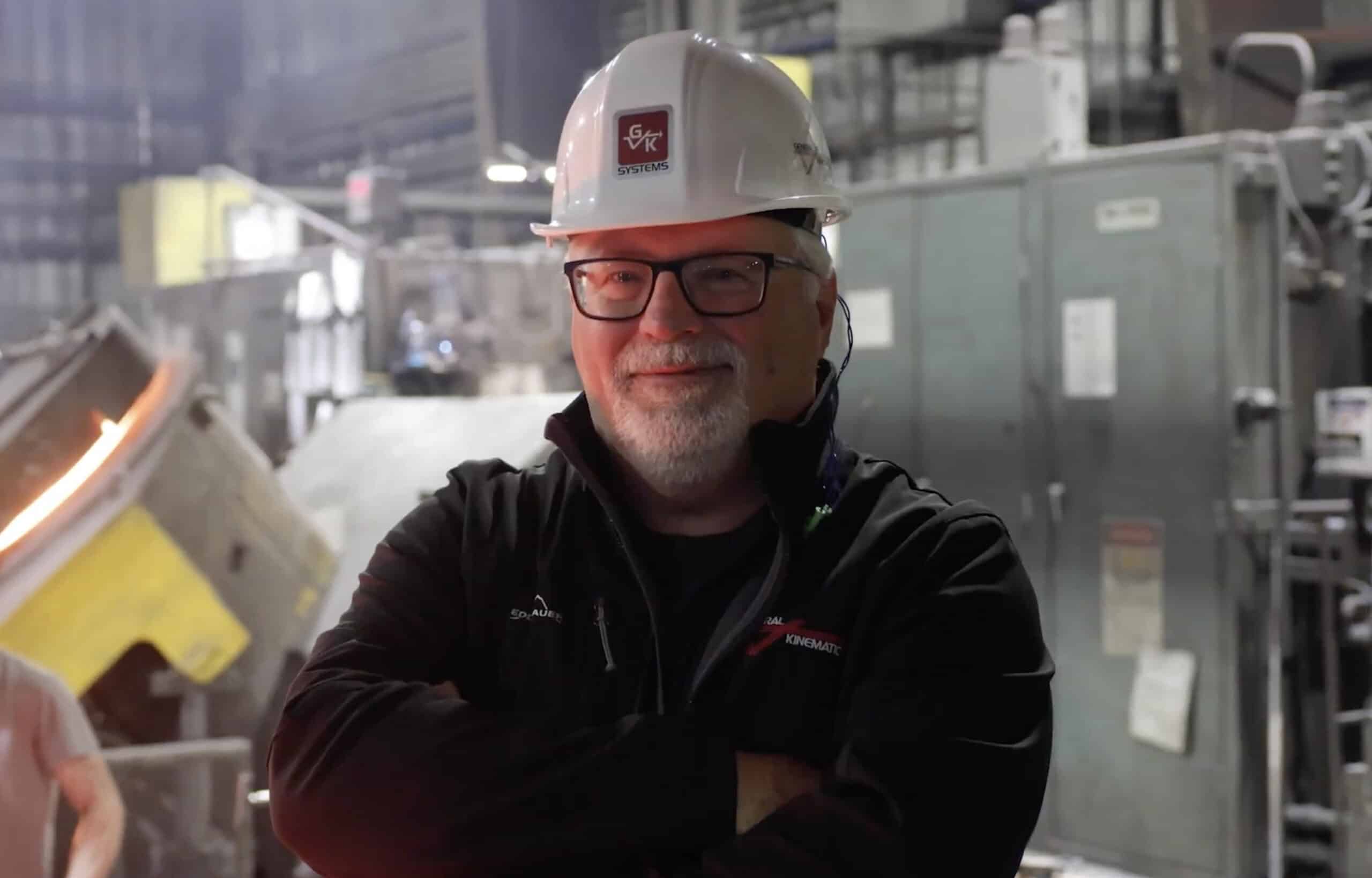A photo of a GK Systems team member smiling at the camera with his arms crossed. He is in a foundry wearing a hat that says "GK SYSTEMS"